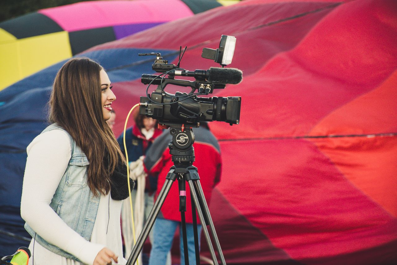 Menu A female videographer operates a camera before a colorful hot air balloon backdrop.