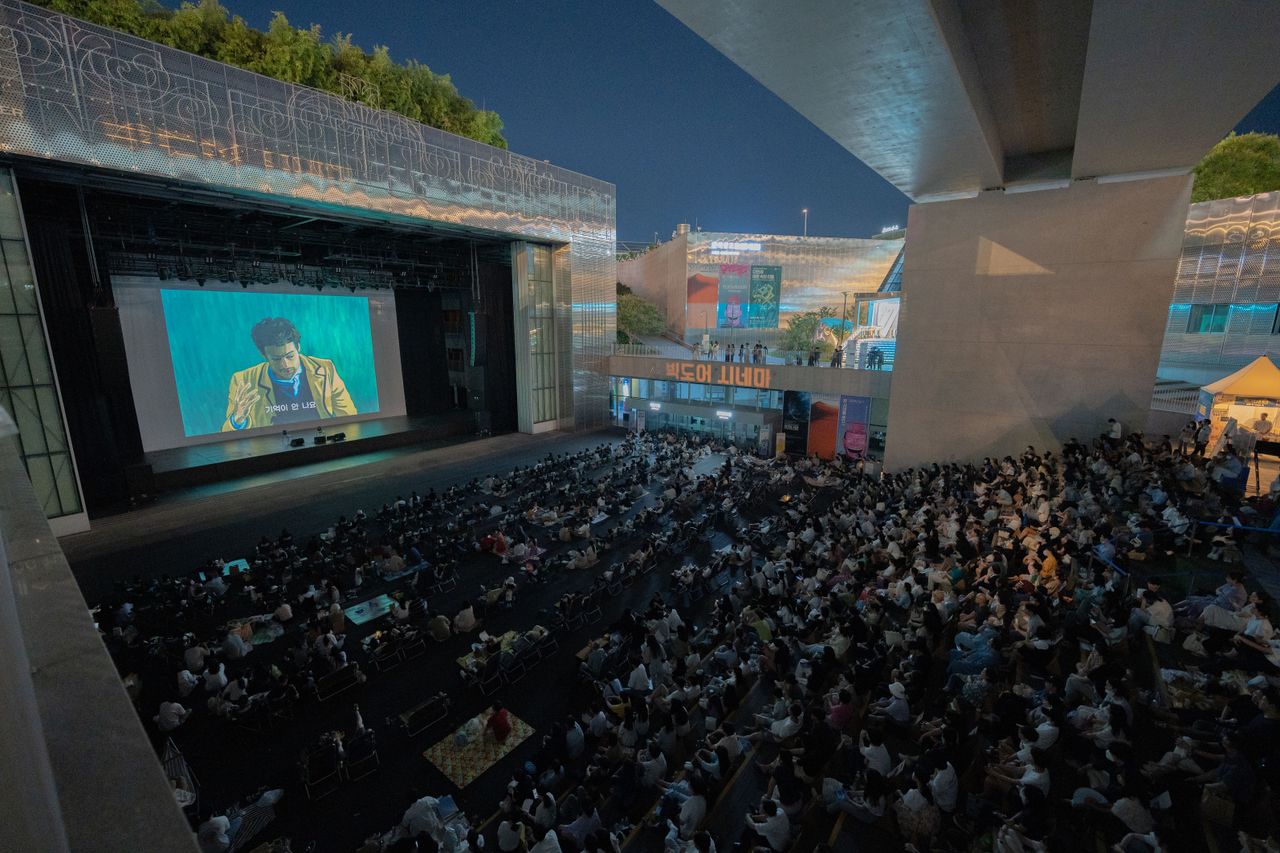 Menu A large audience gathers outdoors at night to watch a movie screening under a bridge.